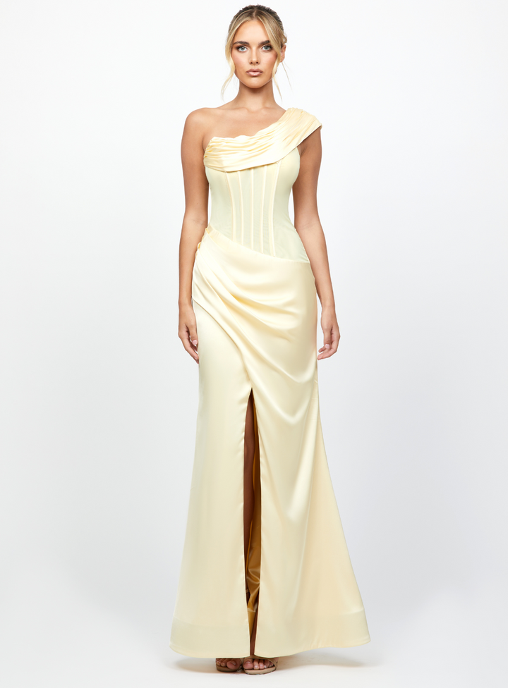 A model wearing a one shoulder, full-length gown with a sheer corset bodice and a thigh-high split, in a lemon yellow color.